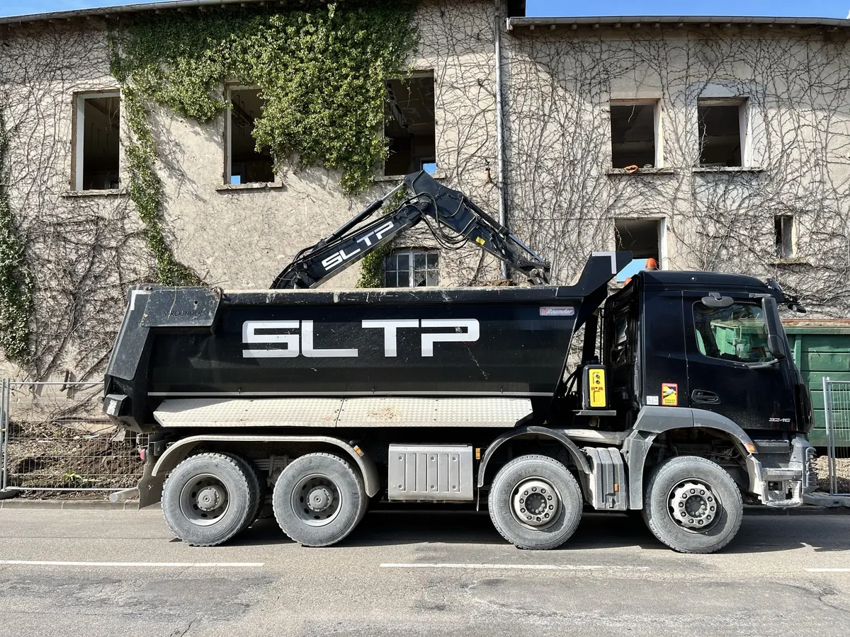 Camion-benne noir siglé SLTP devant un bâtiment couvert de lierre, sur le chantier EPFA, à Chazey-sur-Ain.