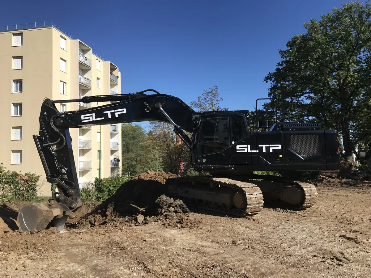 Pelle noire siglée SLTP sur le terrain du chantier Gingko, à Gleizé, devant un immeuble.