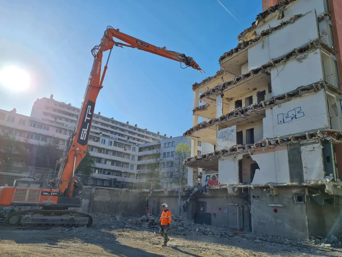 Pelle à grand bras orange contre un immeuble à balcons en démolition, sur le chantier Interface, à Lyon 8.