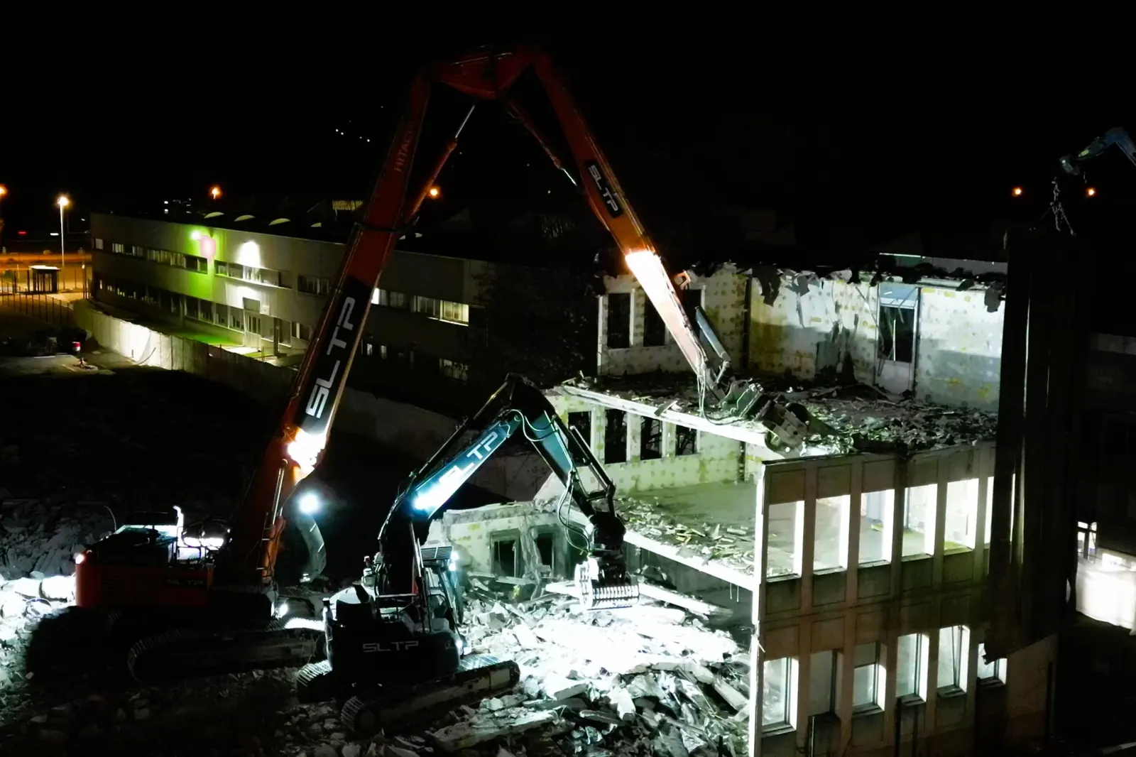 Démolition de nuit sur le chantier Fontanil, près de Grenoble : une pelle à grand bras orange et une pelle noire, siglées SLTP, sous les projecteurs.