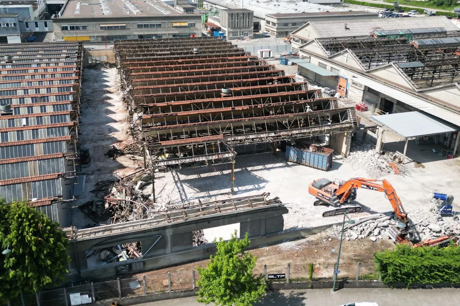Vue aérienne du chantier Vertuose, à Annecy : la charpente d’une grande halle mise à nu, une pelle orange siglée SLTP dans la cour.
